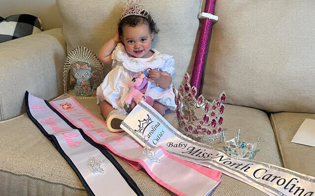 Toddler wearing pageant gown surrounded by tiaras and sashes